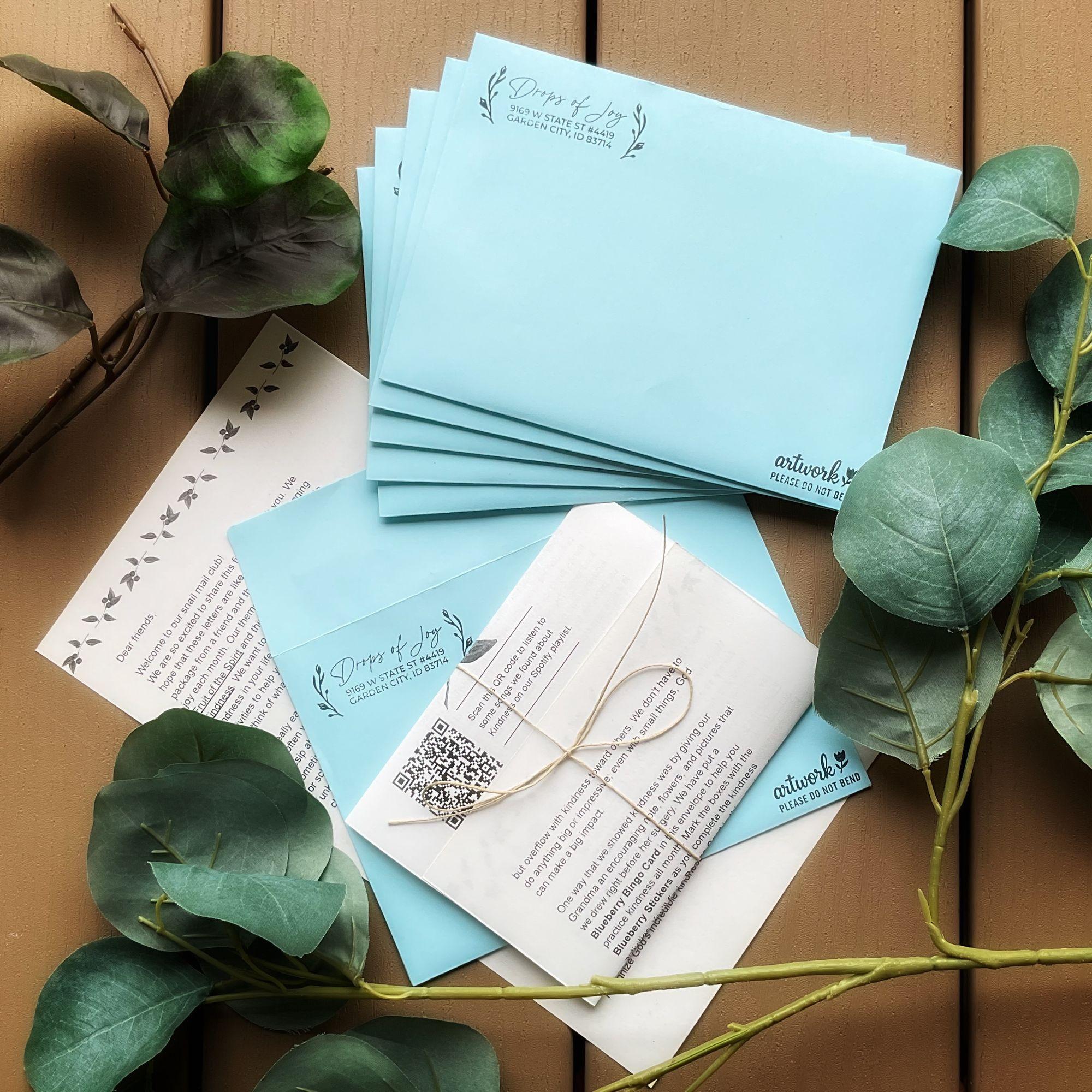 Drops of Joy envelopes, devotional letters, and greenery arranged on a wood backdrop